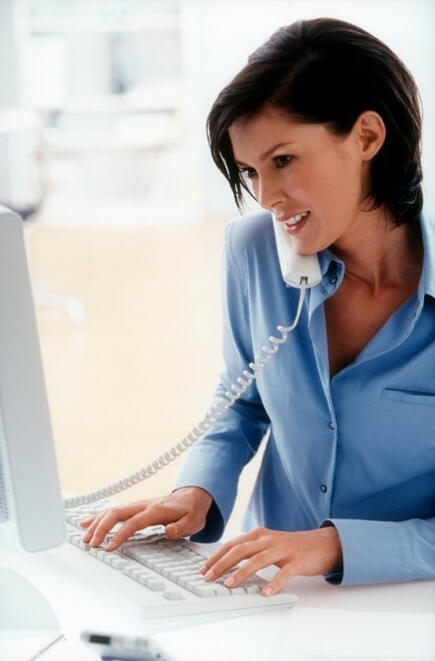 Picture of a dark-haired lady in a blue shirt sitting at a white desk, typing on a white computer keyboard and talking on a white telephone.