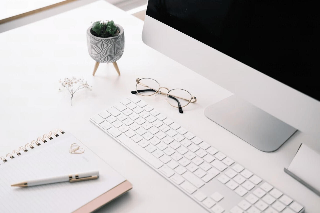 A desk with a keyboard, monitor, glasses, notebook and pen and a plant.