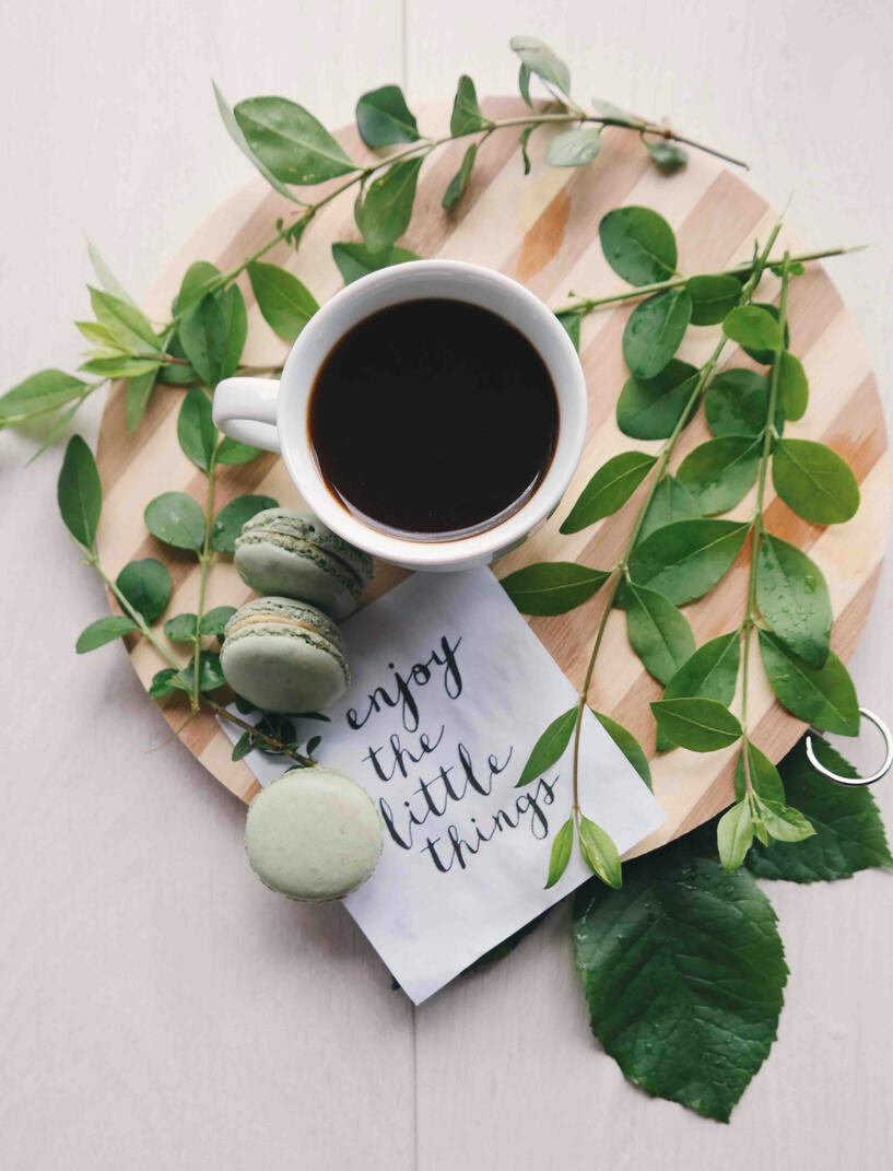 A cup of tea and three mararons on a round, wooden board decorated with greenery and a note that reads, "Enjoy the little things".
